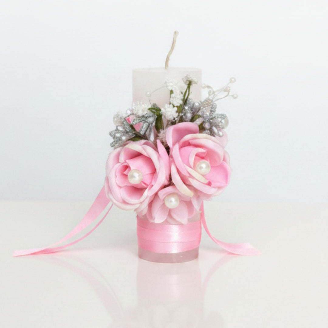 A decorative pink floral candle holder featuring a white pillar candle, pink fabric roses, a pink satin ribbon, and small faux pearls, displayed on a textured brown upholstered chair.