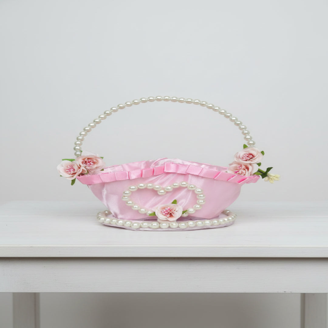 A pink satin basket handbag decorated with faux pearls, a heart-shaped pearl frame, and pink flowers, set against a gray quilted background.