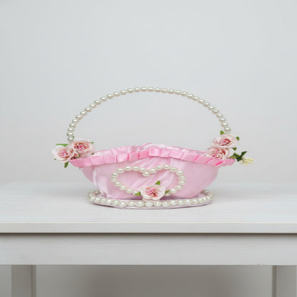 A pink satin basket handbag decorated with faux pearls, a heart-shaped pearl frame, and pink flowers, set against a gray quilted background.